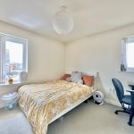 Bright bedroom with bed, desk, and two windows.